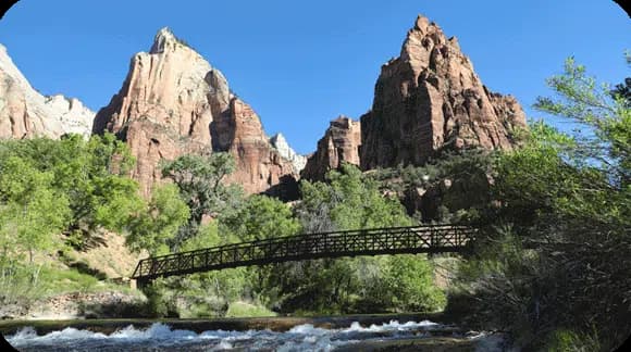 A wooden bridge spans a river, flanked by towering red rock formations and lush greenery under a clear blue sky.
