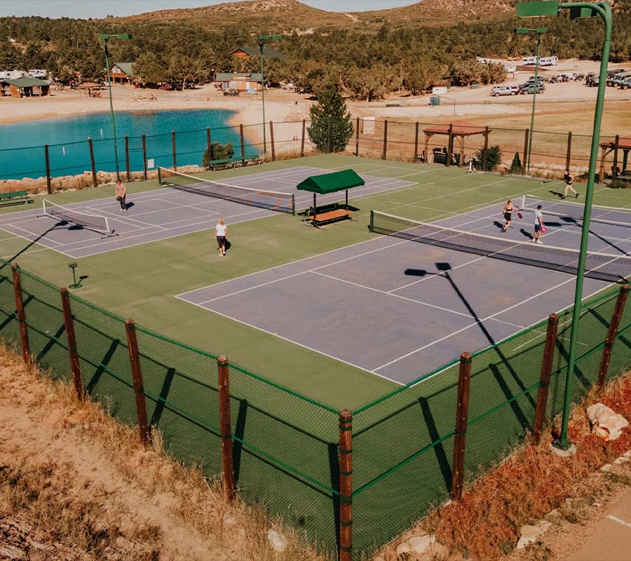 Aerial view of tennis courts with players in action, surrounded by trees and a blue water body in the background.