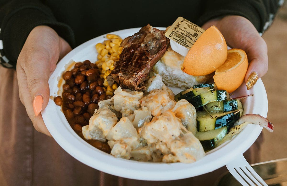 A person holds a plate filled with various foods, including grilled meat, baked beans, corn, zucchini, and fruit.