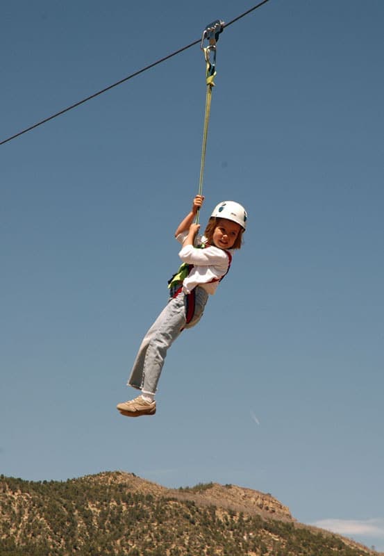 A smiling child rides a zip line against a clear blue sky and mountainous background.