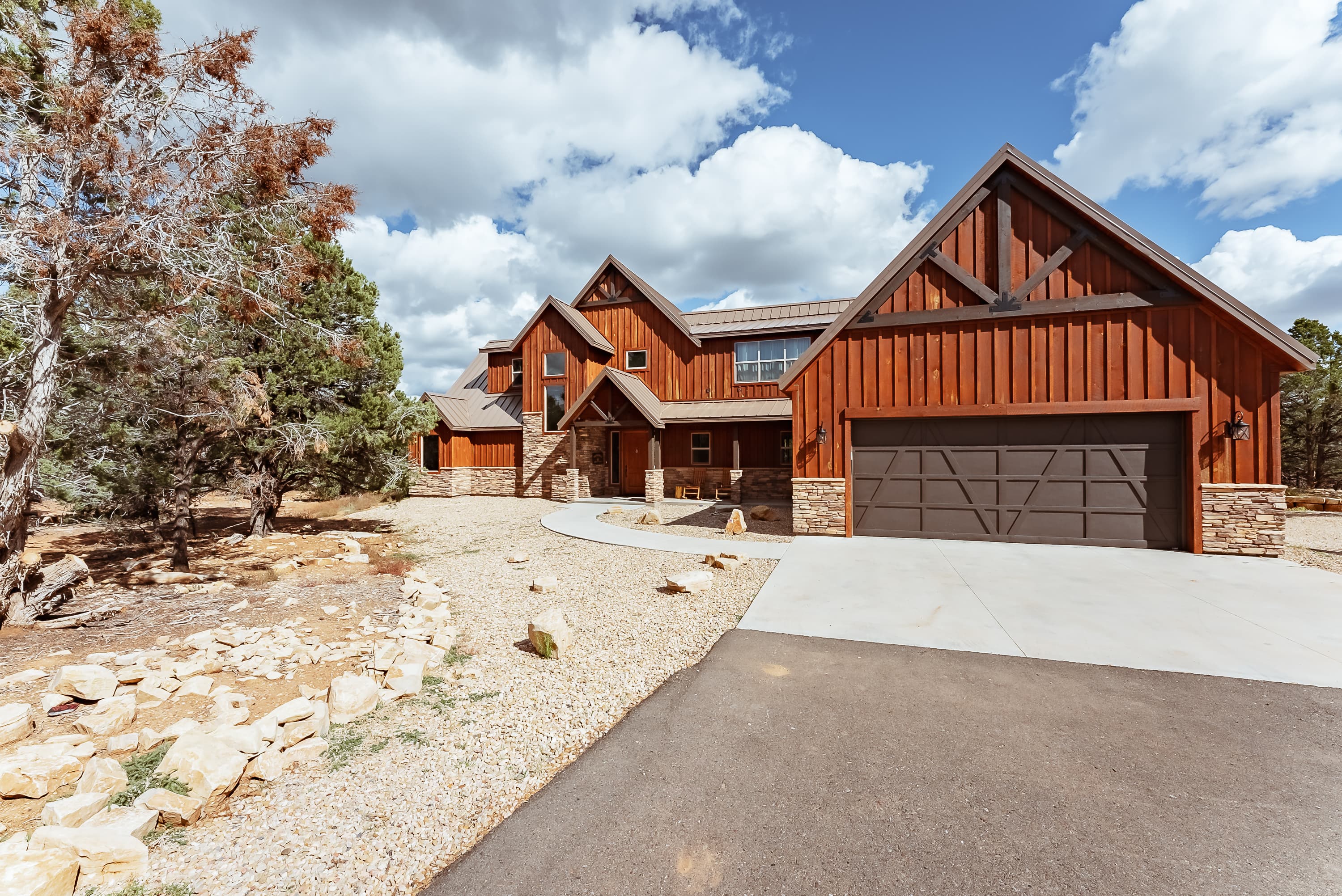 A large wooden house with a stone foundation and a circular driveway, set against a backdrop of blue sky and fluffy clouds.