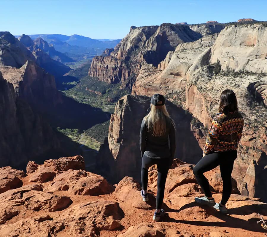 Two women stand on the edge of a canyon overlooking a vast landscape.