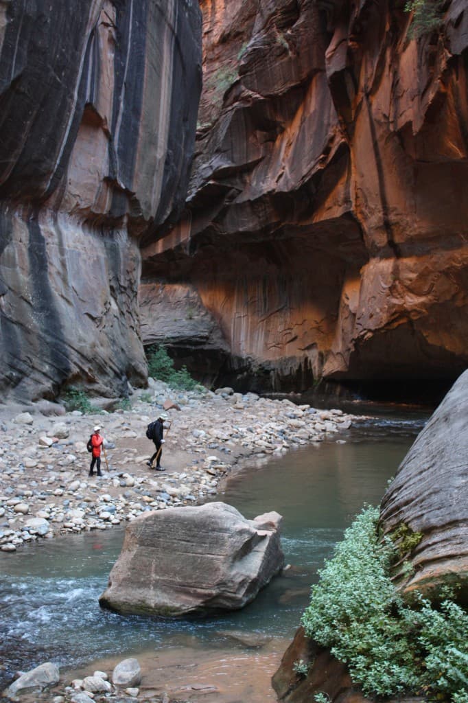 Two hikers explore a narrow canyon with a winding stream and steep rock walls.