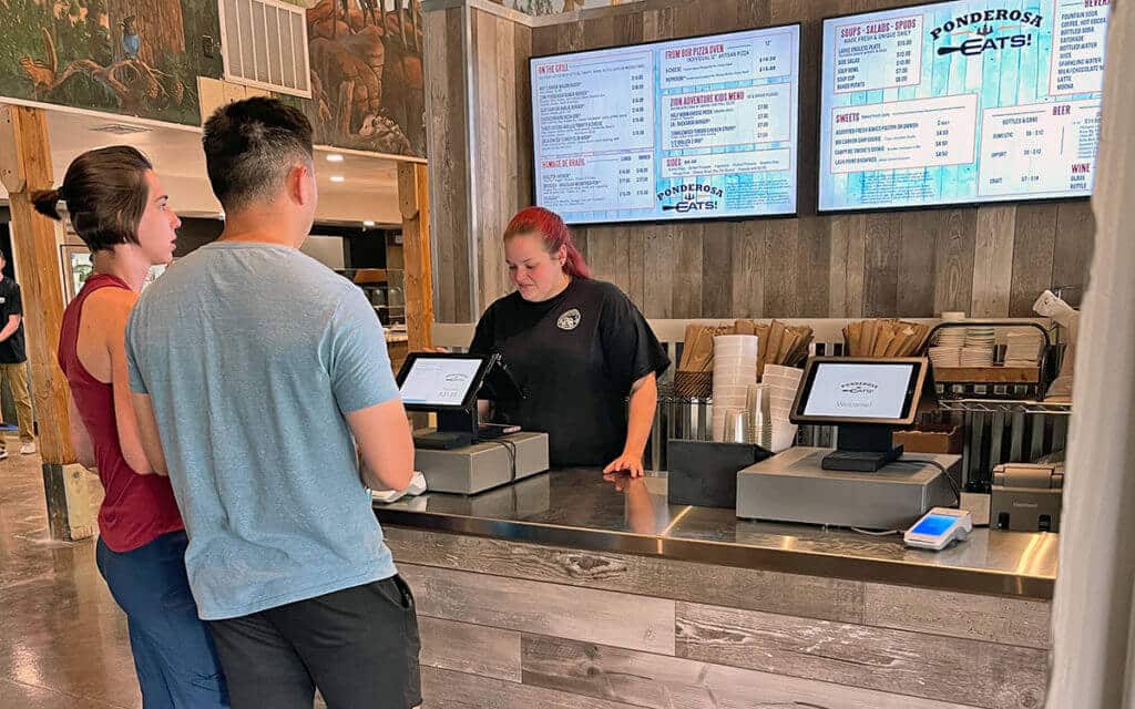 Two customers interact with a cashier at a restaurant counter displaying menus on screens.