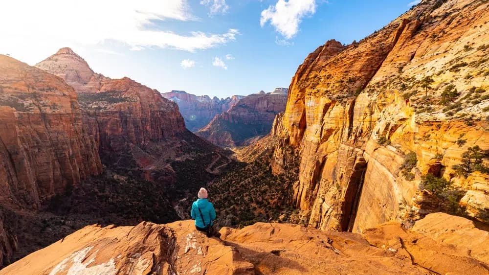 A person in a blue jacket sits on a rocky ledge overlooking a vast canyon landscape.