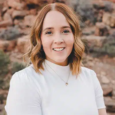 A woman with wavy, shoulder-length hair smiles warmly against a rocky backdrop.