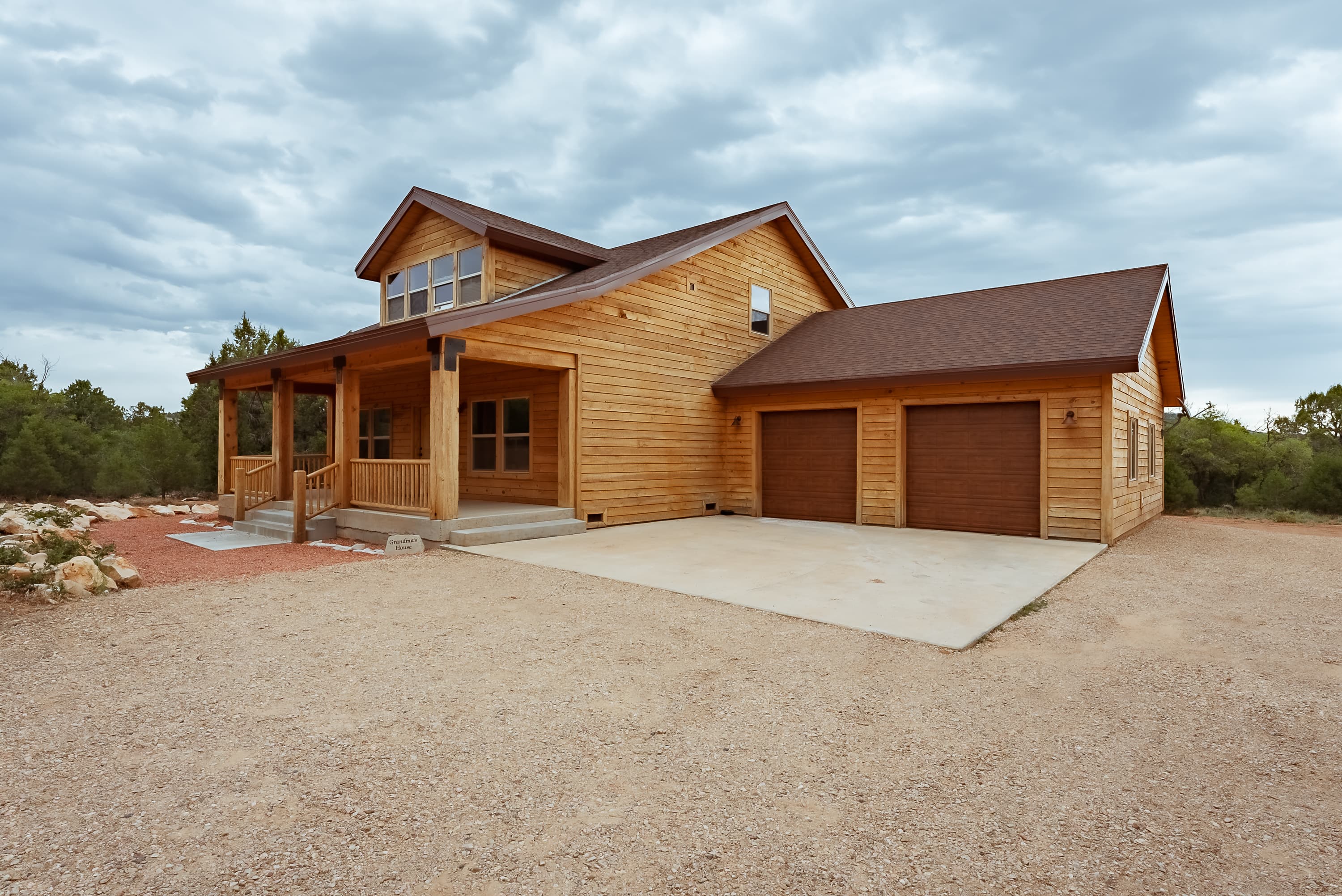 A wooden two-story house with a front porch and a two-car garage, surrounded by gravel and sparse landscaping under a cloudy sky.