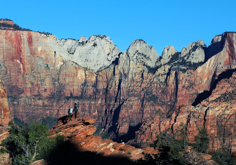Hikers stand on a rocky outcrop overlooking the towering cliffs of Zion National Park under a clear blue sky.