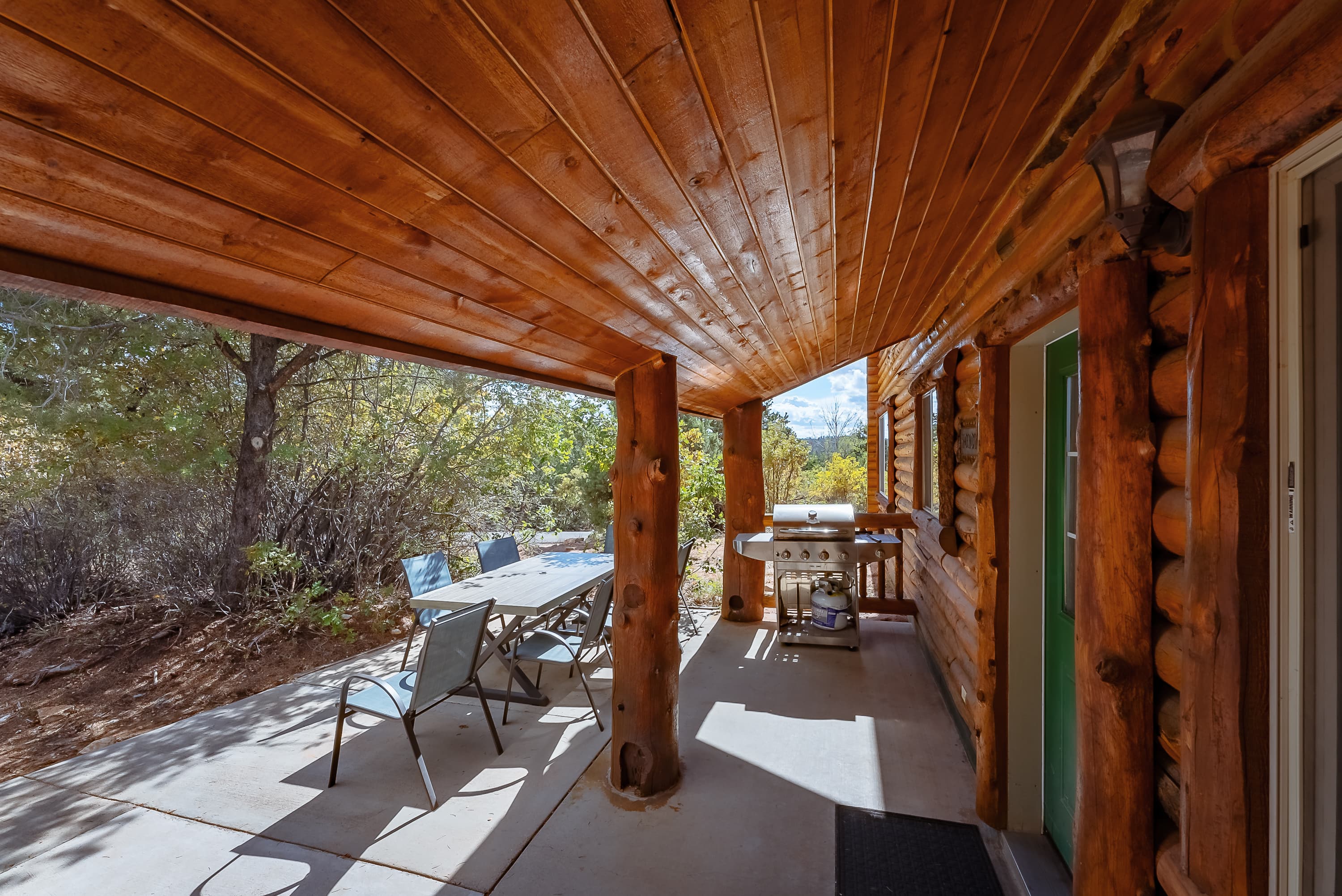 A rustic porch with a wooden ceiling, a table and chairs, and a grill, surrounded by trees.