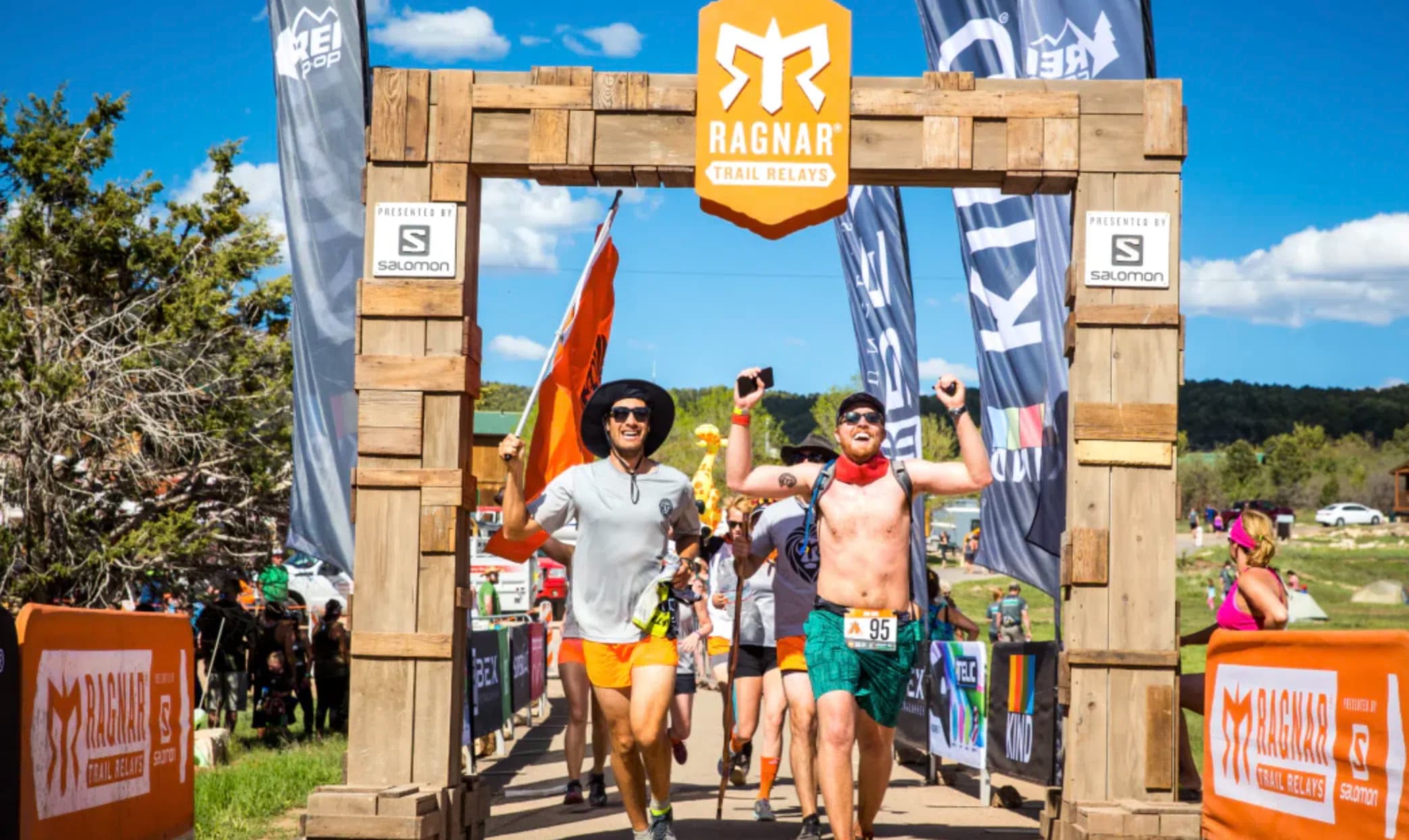 Two runners joyfully cross a wooden finish line adorned with "Ragnar" branding at a trail running event.