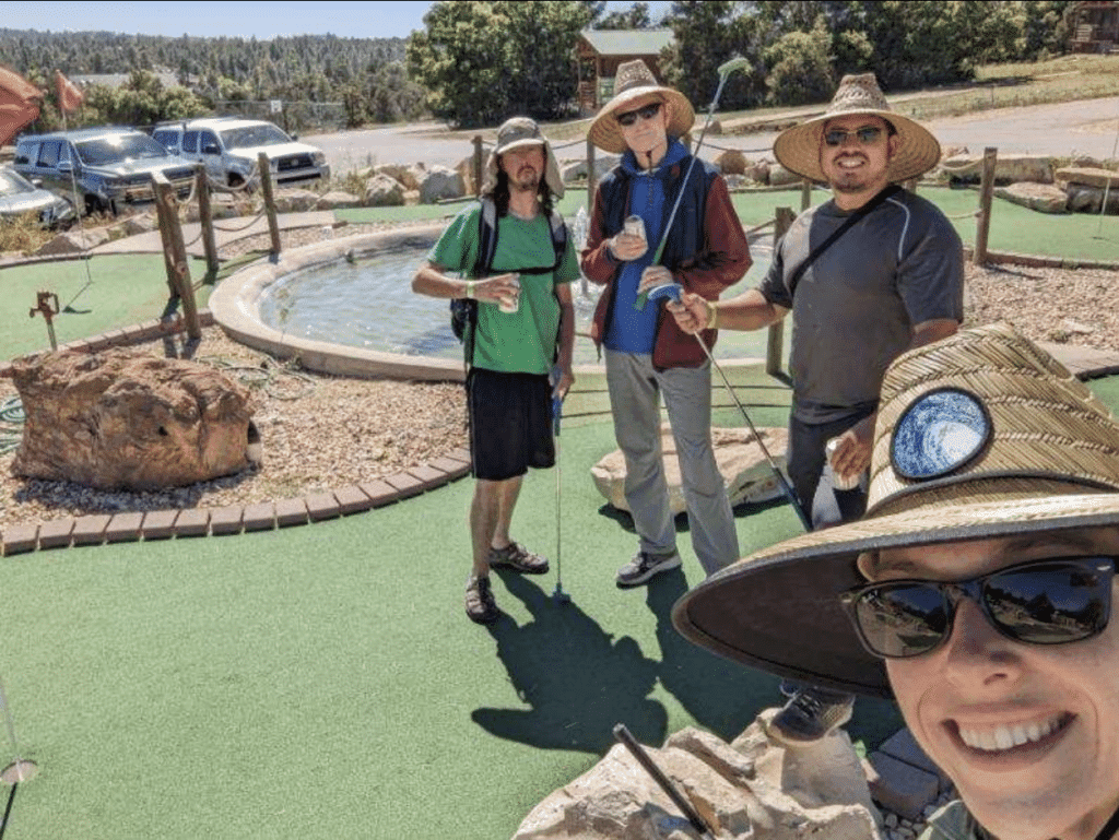 Four friends pose together at a mini-golf course, surrounded by greenery and a small pond.