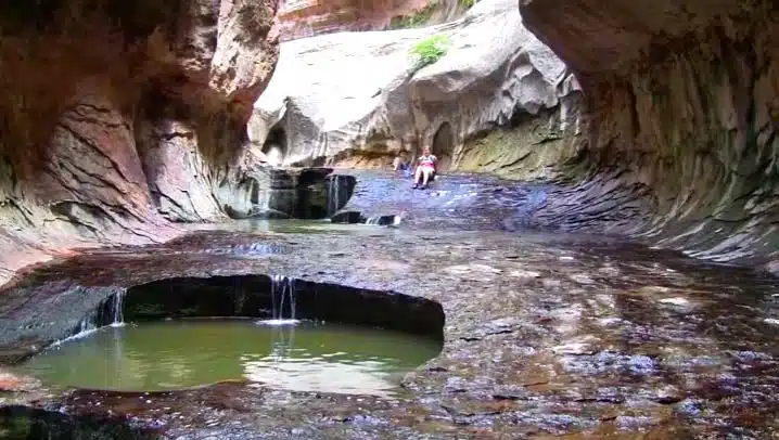 A person sits on a rocky ledge near a shallow pool in a gorge with smooth, curved walls.
