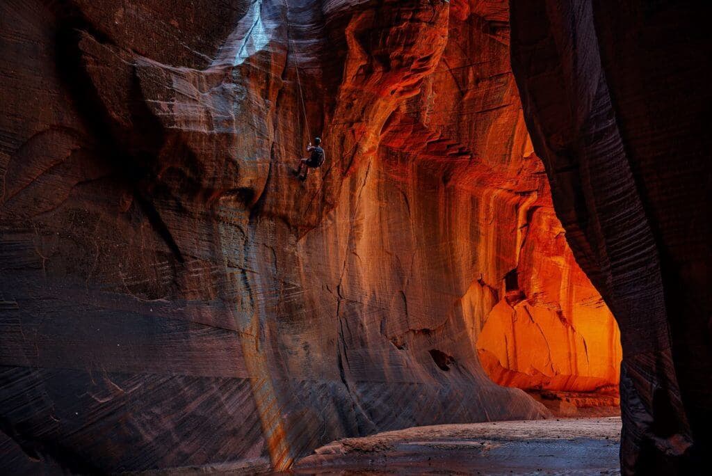 A climber scales a vibrant red sandstone canyon wall illuminated by warm sunlight.