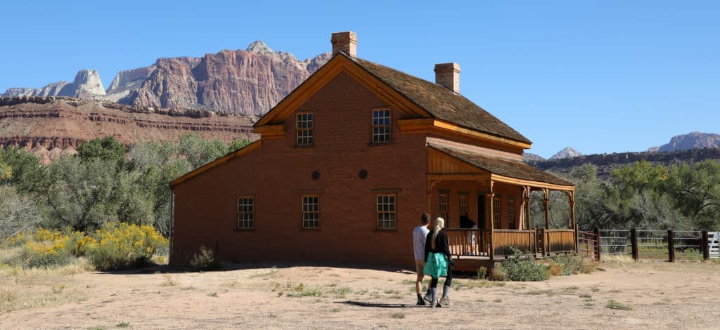 A couple walks towards a rustic brick house with mountains in the background.