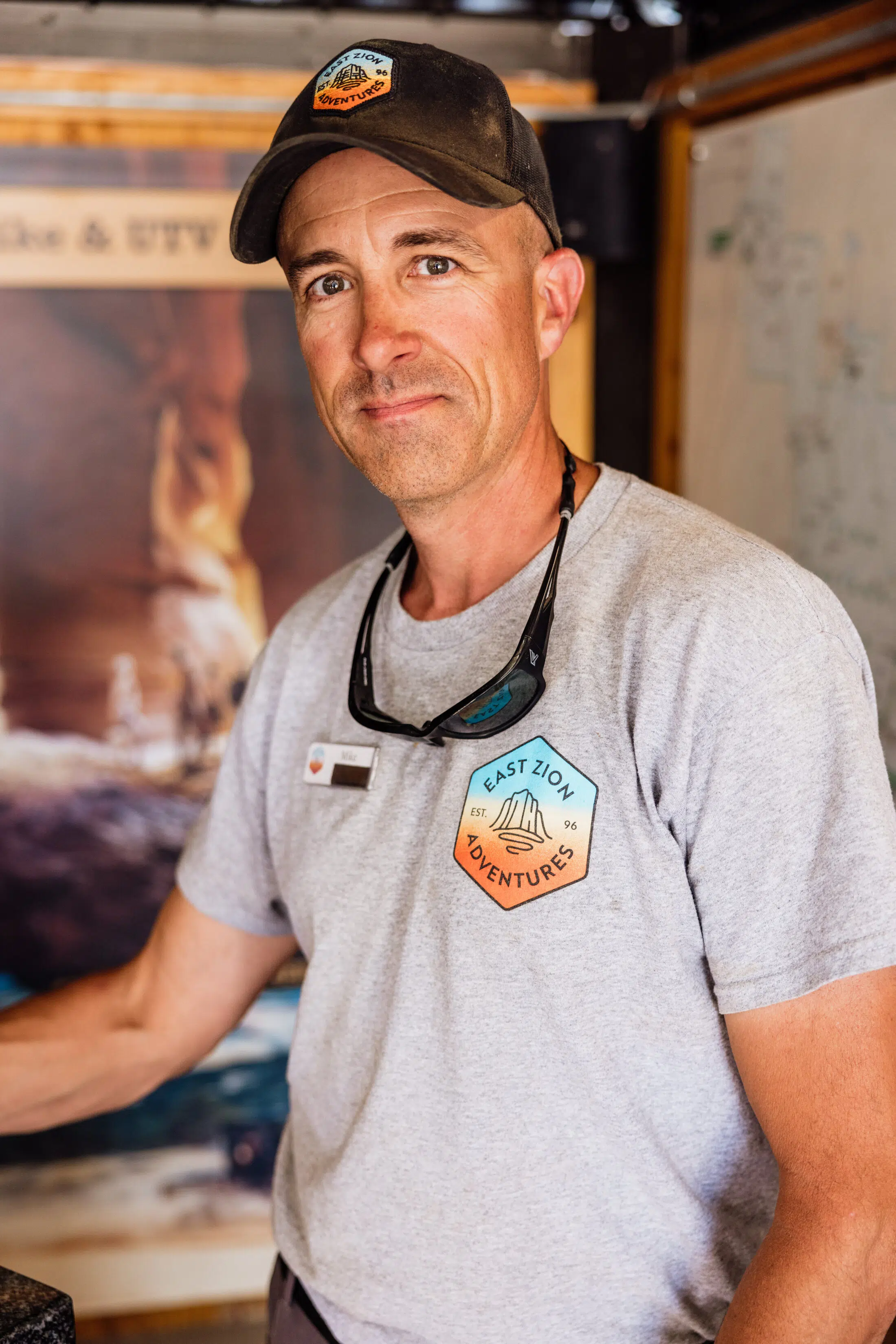 A man wearing a cap and a gray t-shirt with the logo "East Zion Adventures" smiles in an indoor setting.