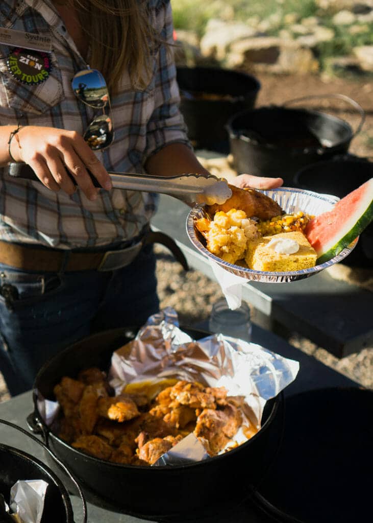 A person is serving food from a pot using tongs, filling a plate with fried chicken, corn, and watermelon.