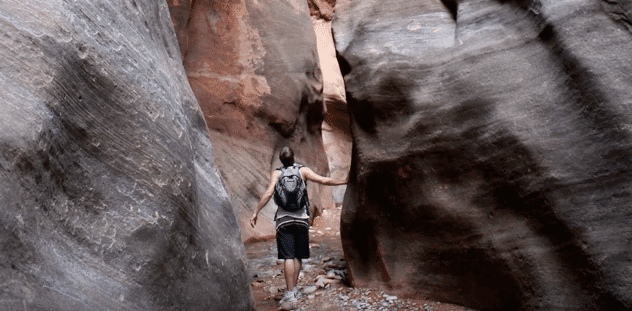A person with a backpack walks through a narrow canyon surrounded by smooth, rocky walls.