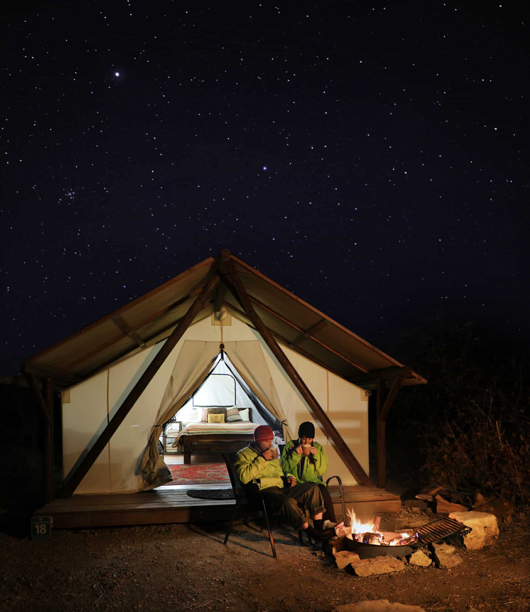 Two people sit by a campfire in front of a glamping tent under a starry night sky.