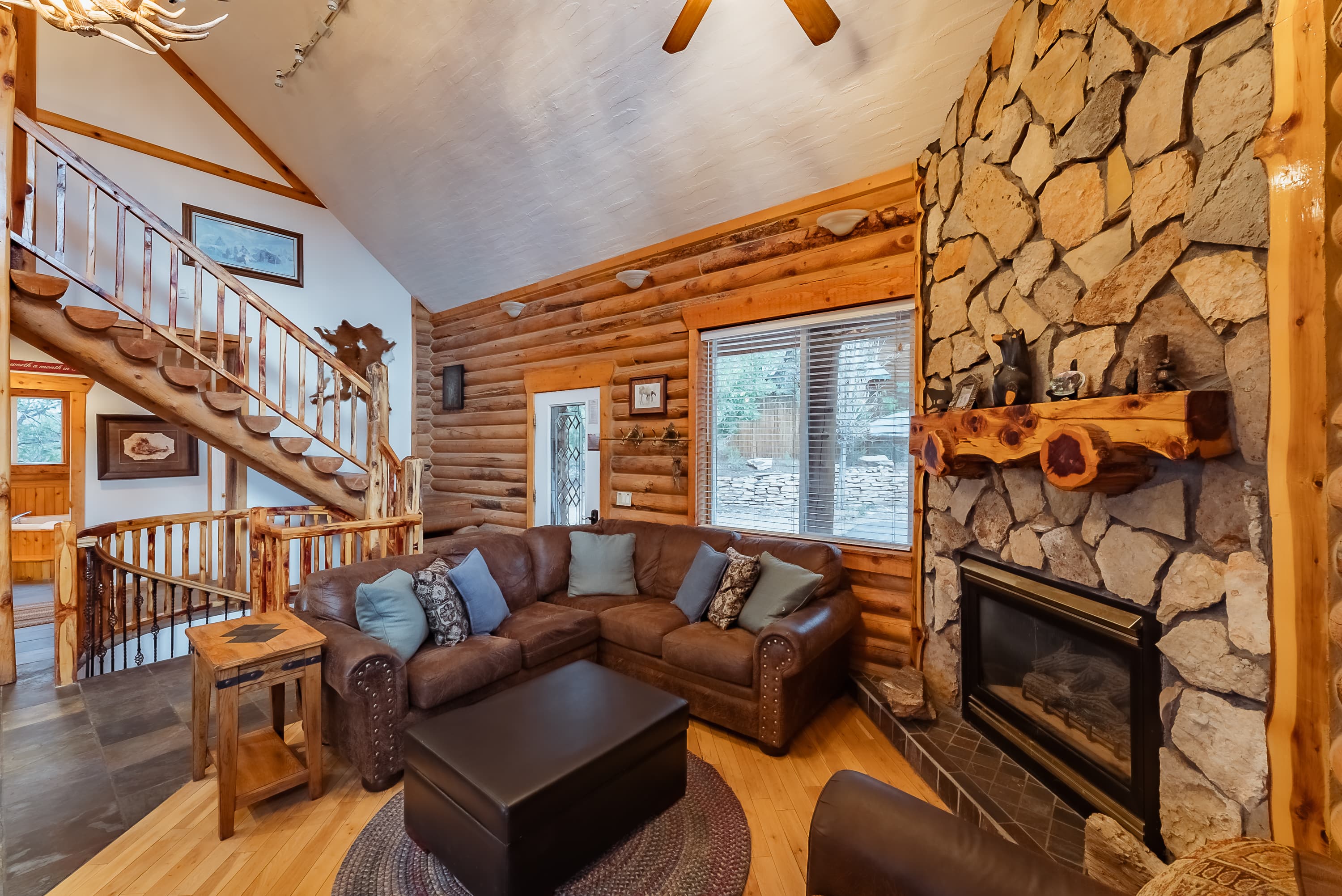 Cozy living room with a stone fireplace, leather furniture, and wooden staircase in a log cabin.