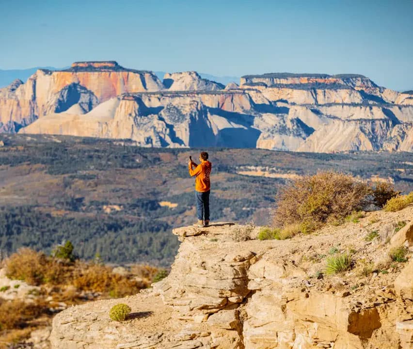 A person in an orange jacket stands on a rocky ledge, taking a photo of a mountainous landscape.