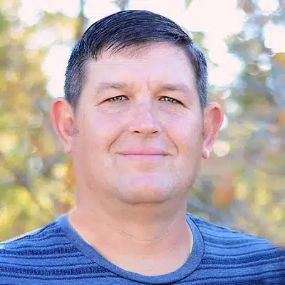 A smiling man with short dark hair, wearing a striped blue shirt, is posing outdoors.
