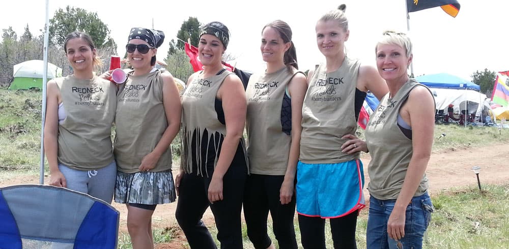 A group of six women wearing matching tan tank tops poses together outdoors at a casual gathering.