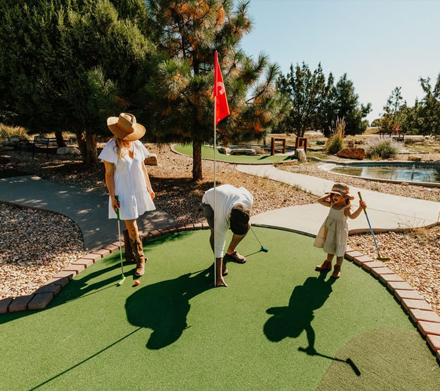 A family enjoys mini-golf on a sunny day, with a woman and child holding putters while a man retrieves a golf ball.