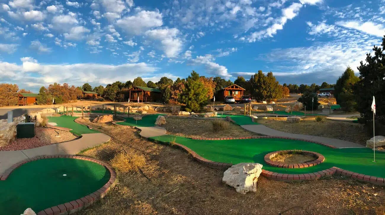 A scenic mini-golf course with lush green turf, surrounded by trees and a blue sky.