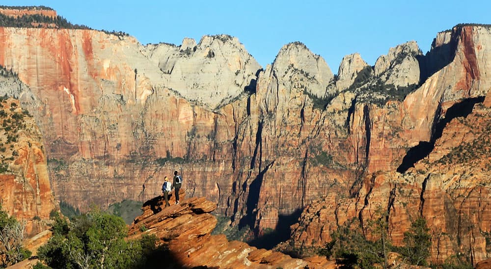 Three hikers stand on a rock ledge with dramatic red rock formations in the background.