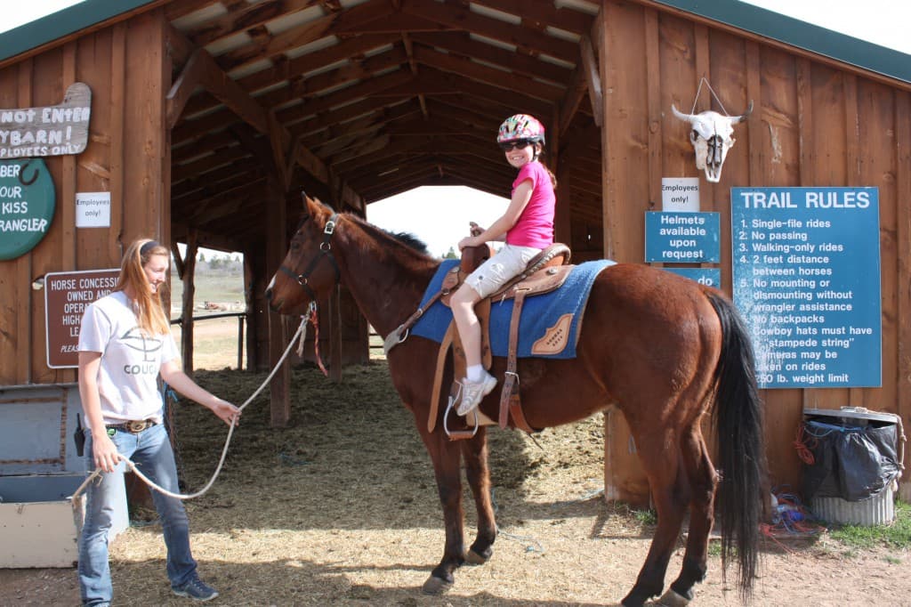 A girl wearing a helmet sits on a horse while a person holds its reins at a stable.