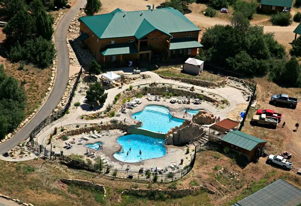 Aerial view of a large, landscaped pool area alongside a wooden house and surrounding greenery.
