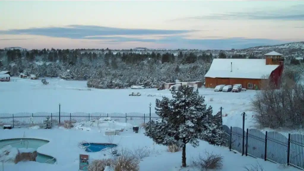 A snowy landscape featuring a red barn, pine trees, and a fenced yard.