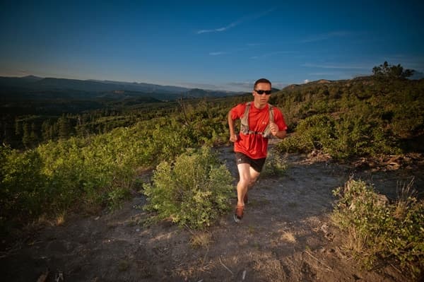 A runner in a red shirt jogs along a trail with a scenic mountainous backdrop.