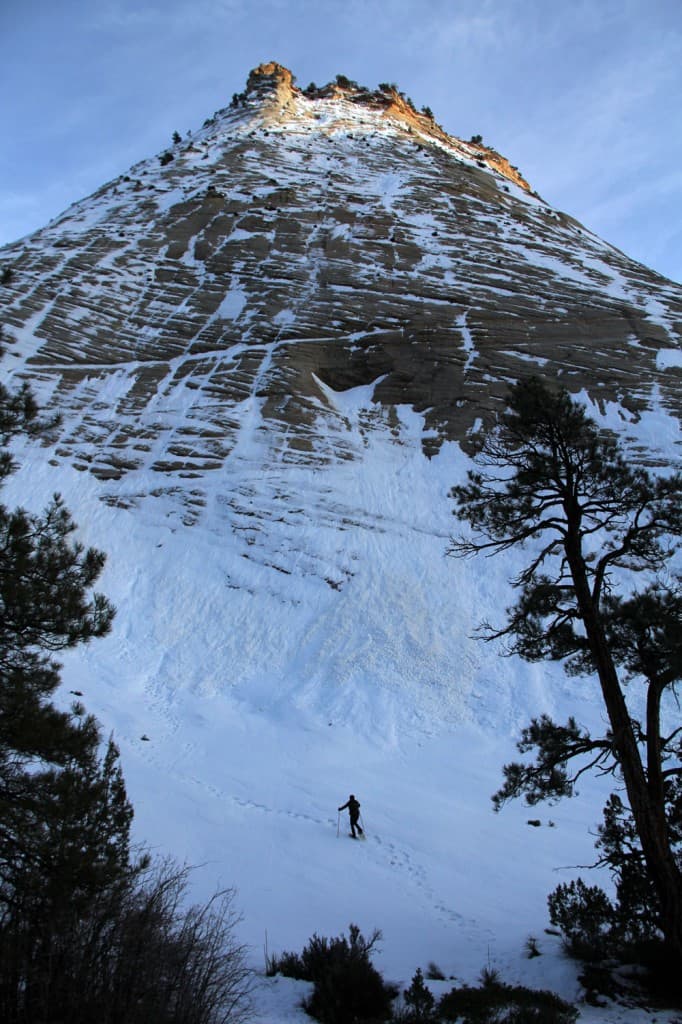 A lone figure hikes up a snowy slope beneath a towering, rocky peak.
