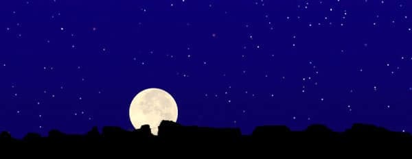A full moon rises above a dark silhouette of mountains against a starry night sky.
