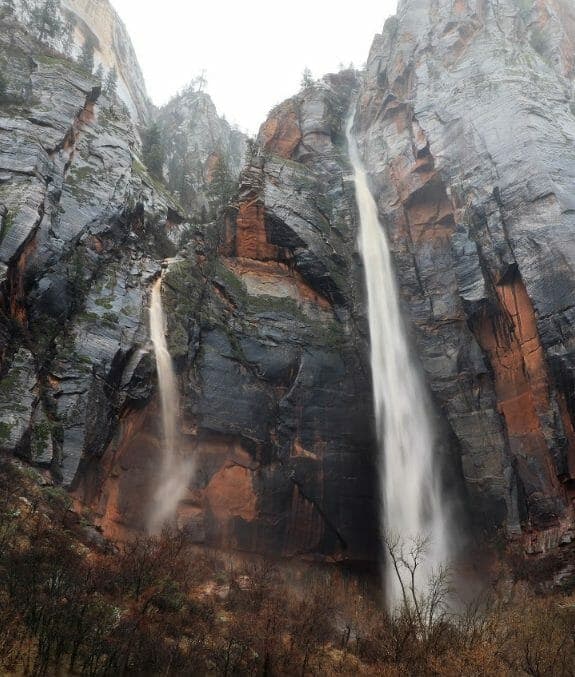 Two waterfalls cascade down a rocky cliff face surrounded by evergreens in a misty landscape.
