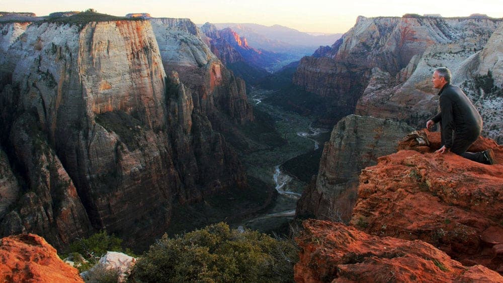 A person kneels on a rocky ledge overlooking a vast canyon at sunset.