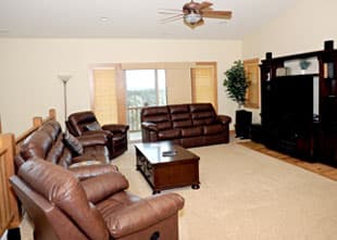 A spacious living room with brown leather sofas, a coffee table, and a television unit, featuring large windows and light-colored walls.