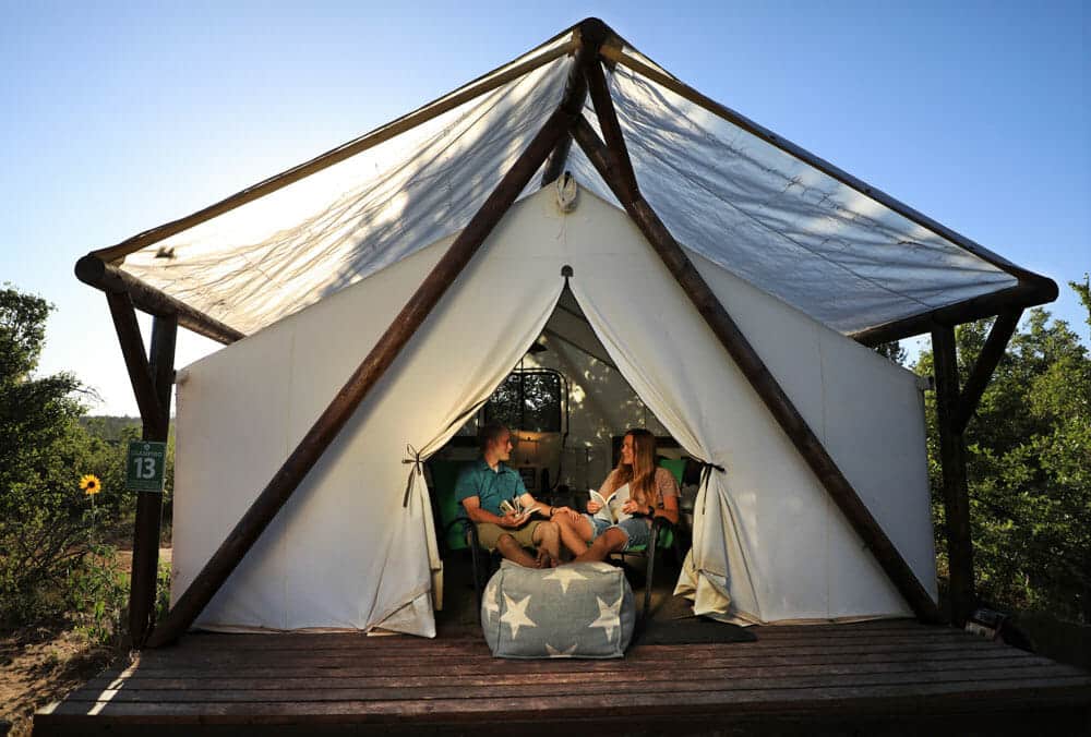 A couple relaxes inside a tent-like structure on a wooden deck, surrounded by nature.