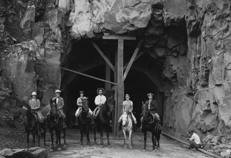 A group of six people on horseback stands in front of a rocky mine entrance.