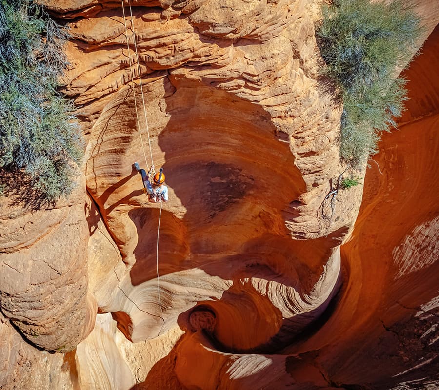 Two climbers rappel down into a steep, winding canyon with reddish rock formations.