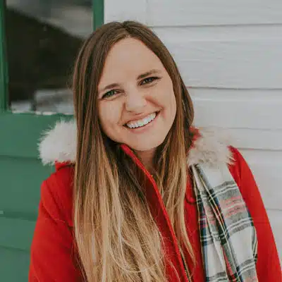A smiling woman with long hair wearing a red coat and a plaid scarf stands in front of a light-colored wall.