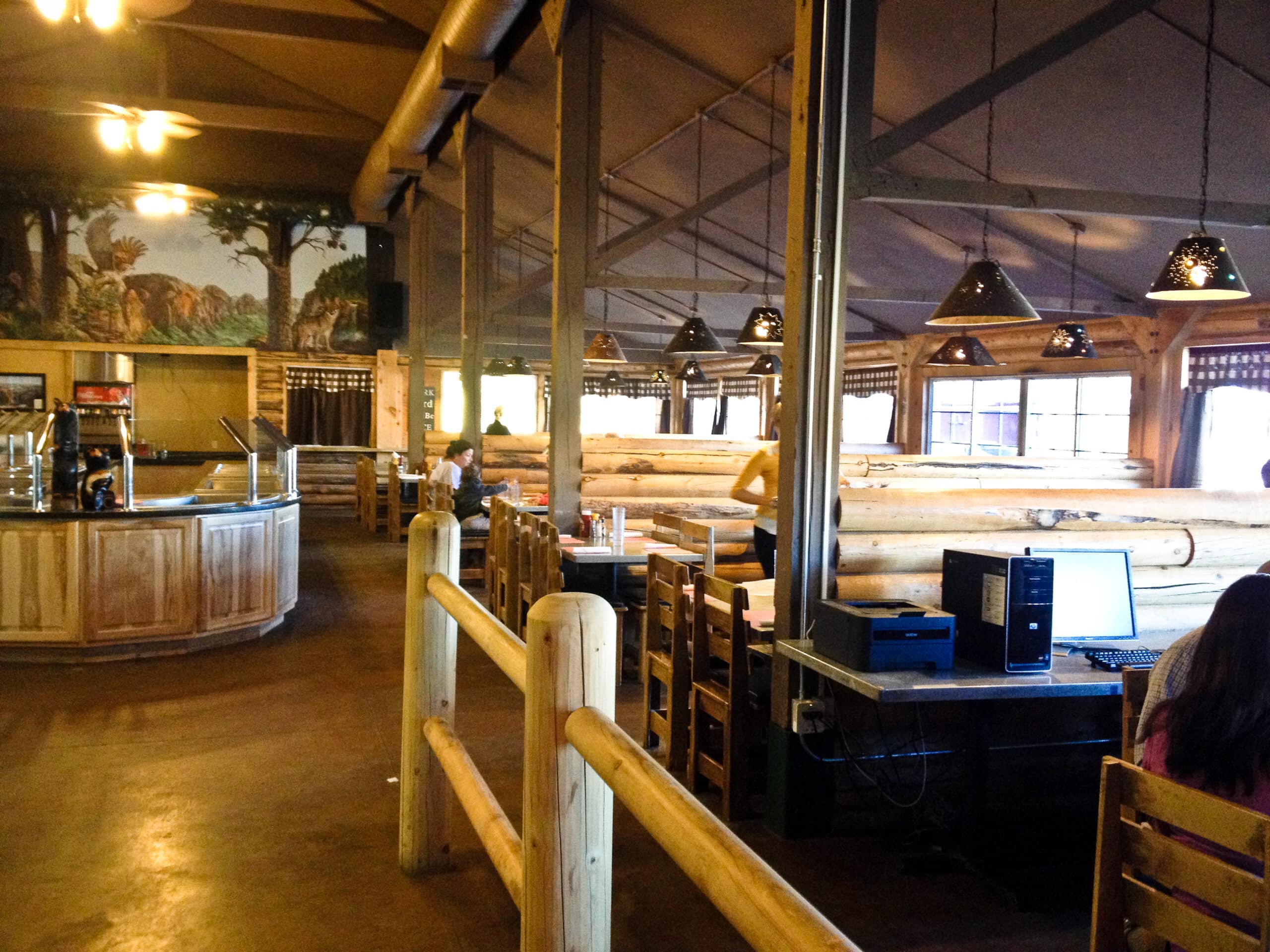 Interior of a rustic restaurant featuring wooden furnishings and a counter, with a few patrons seated at tables.