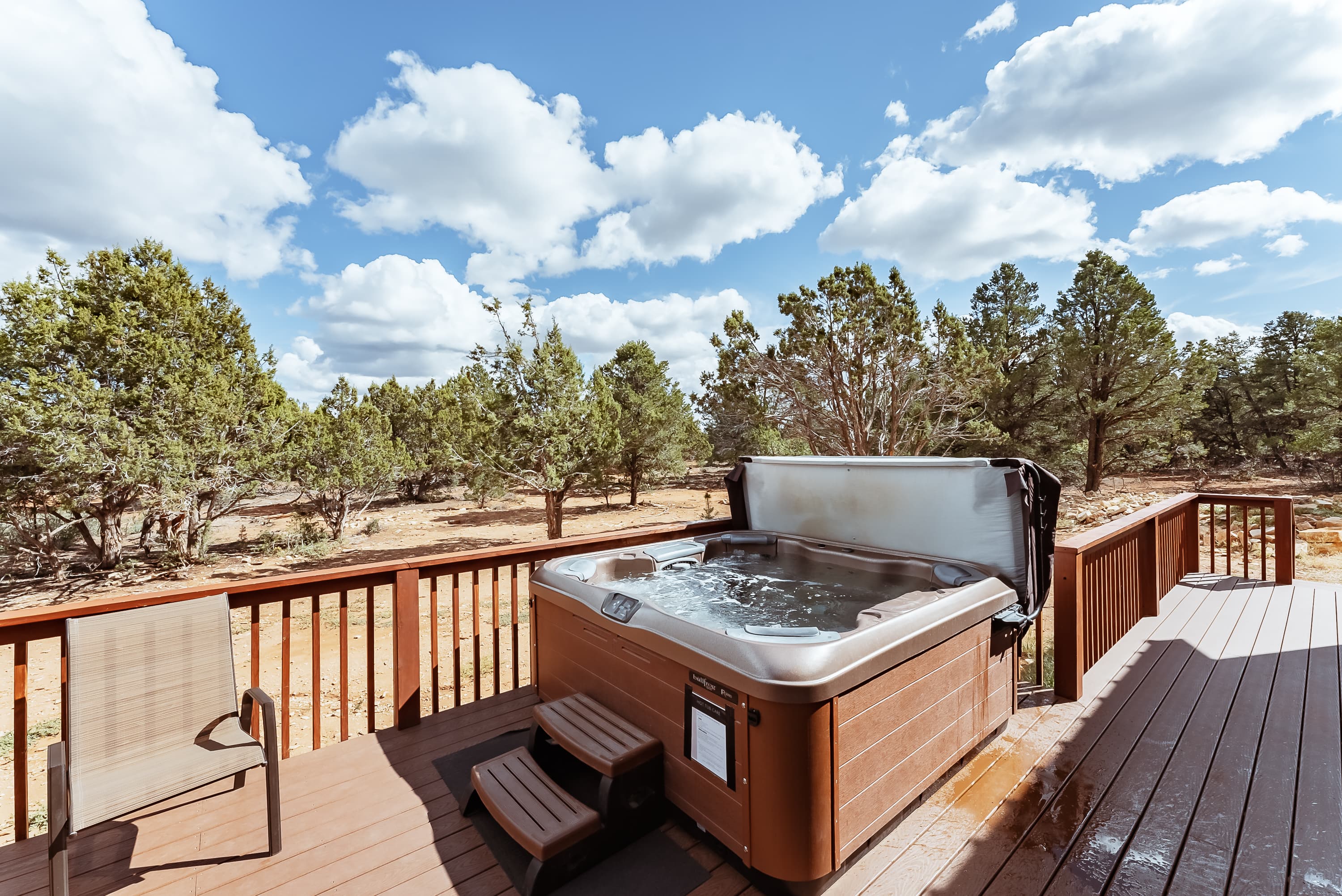 A hot tub on a wooden deck surrounded by trees and a blue sky with fluffy clouds.