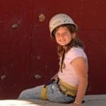 A girl in a climbing helmet sits on a ledge near a rock wall, smiling confidently.