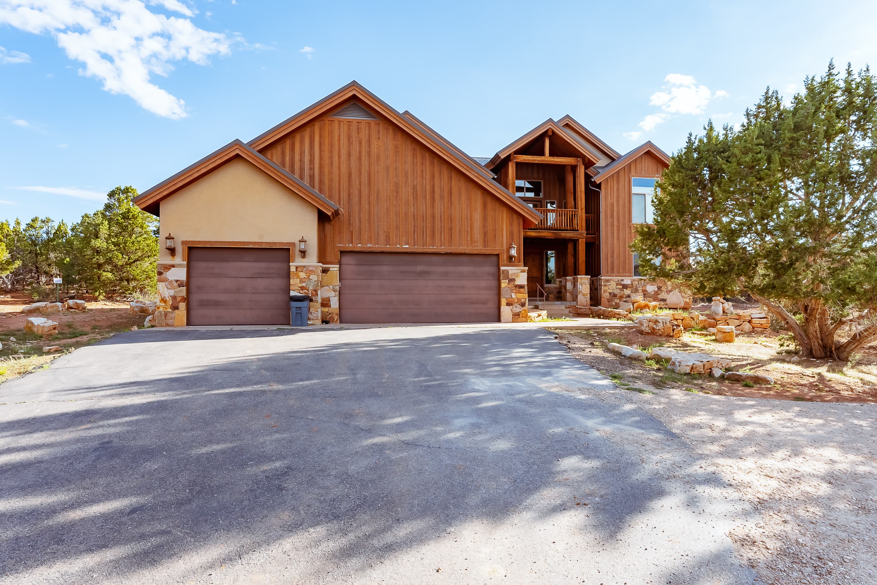 A large, modern house with a stone and wood exterior, featuring a two-car garage and surrounded by trees.