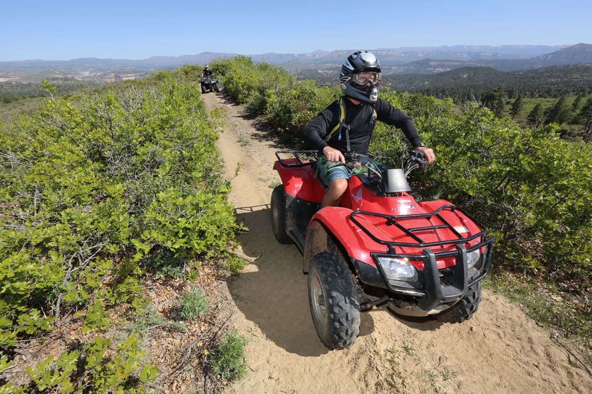 A person riding a red ATV on a sandy trail surrounded by greenery and mountains.