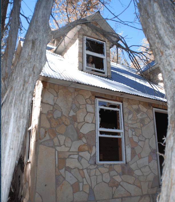 A person is peering out from a broken window of an old stone house framed by trees.