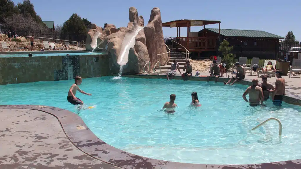 A group of people enjoy a sunny day at a pool with a water slide and rock formations.