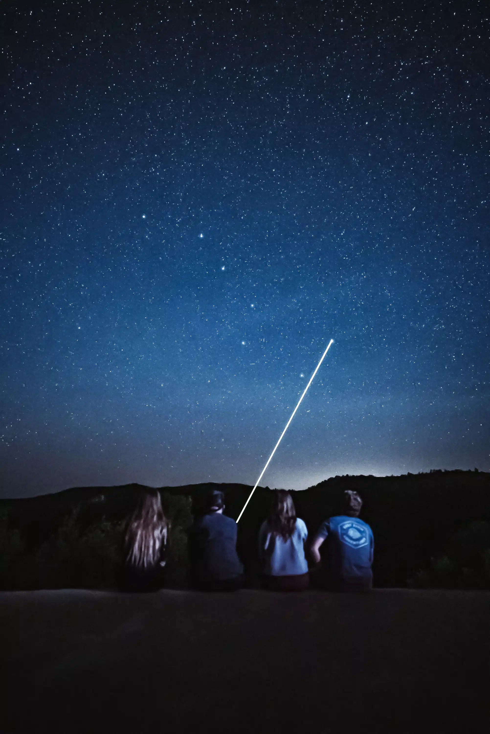 Four people sit on a ledge gazing up at a star-filled night sky with a bright light streaking across.